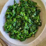 bowl of cooked spinach with minced garlic, olive oil and garlic cloves in the background