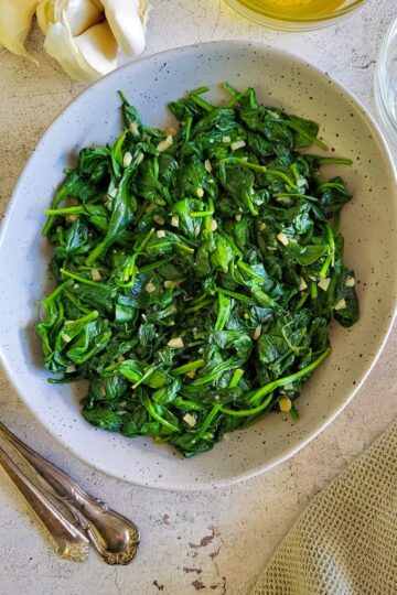 bowl of cooked spinach with minced garlic, olive oil and garlic cloves in the background