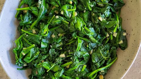 bowl of cooked spinach with minced garlic, olive oil and garlic cloves in the background