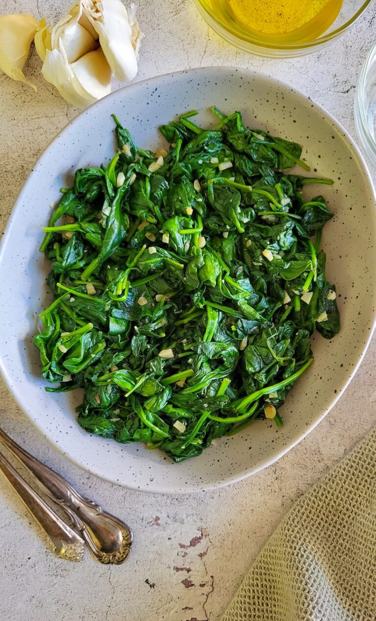 bowl of cooked spinach with minced garlic, olive oil and garlic cloves in the background