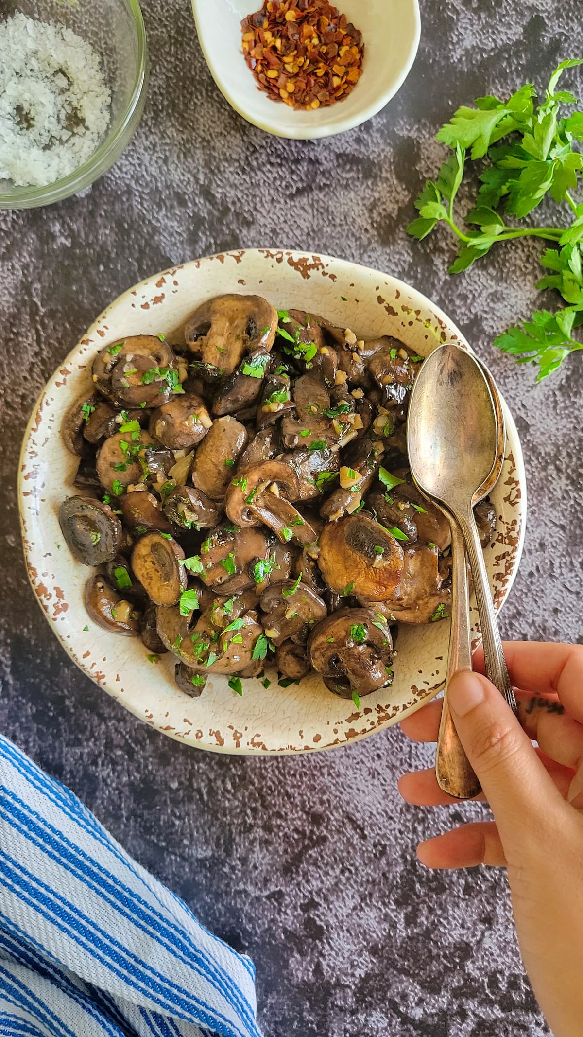cooked sliced mushrooms with garlic and chopped parsley in a bowl with two spoons, hand on the spoons, chili flakes, salt and herbs in the background