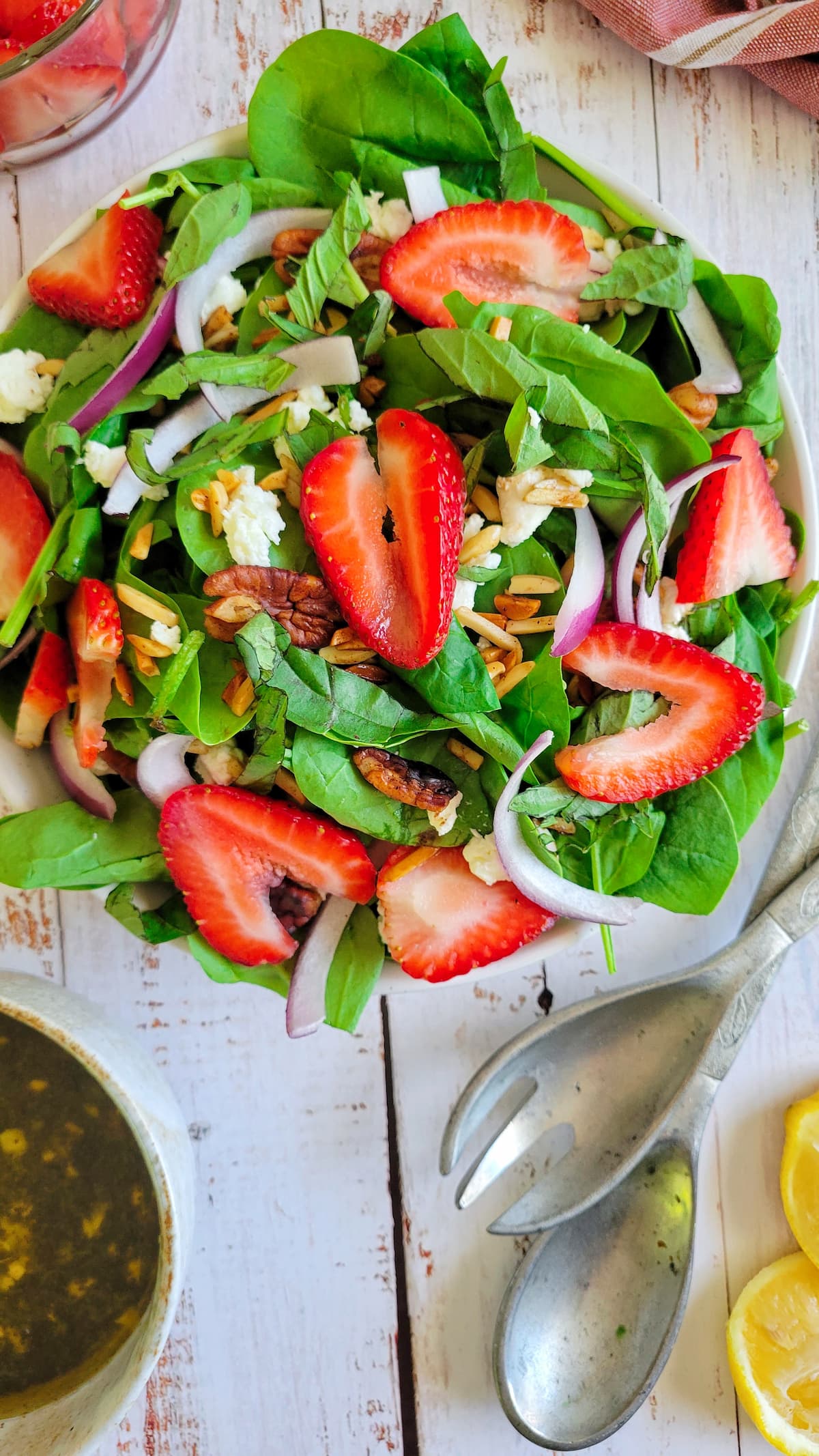 salad with spinach, sliced strawberries, goat cheese, red onions and pecans, next to serving utensils, bowl of dressing and lemon