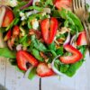 two forks on a bowl of salad with spinach, sliced strawberries, goat cheese, red onions and pecans, bowl of sliced strawberries in the background