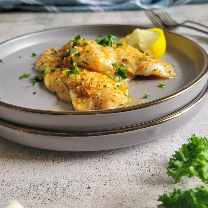 two crusted parmesan fish pieces on a double plate, garnished with fresh chopped parsley and a lemon wedge
