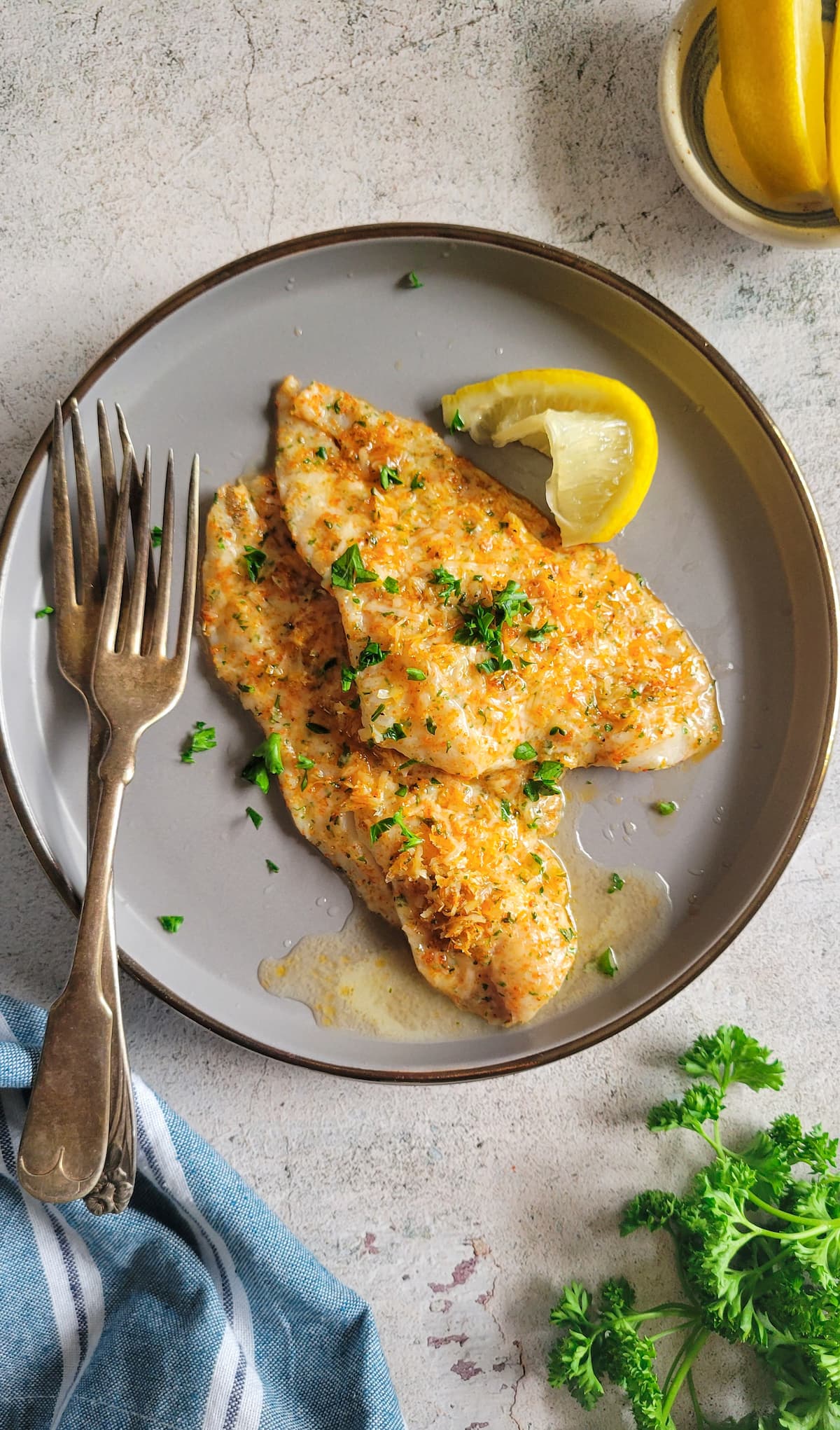 two forks on a plate with two fish fillets crusted in parmesan, fresh chopped parsley and a lemon wedge