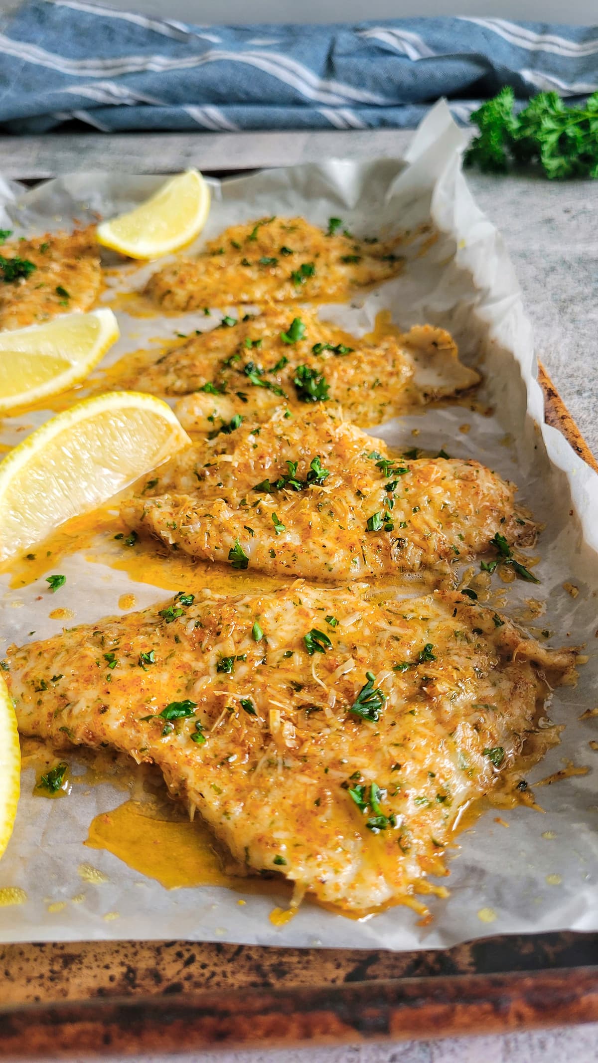 sheet pan of parmesan crusted fish garnished with fresh chopped parsley and lemon wedges