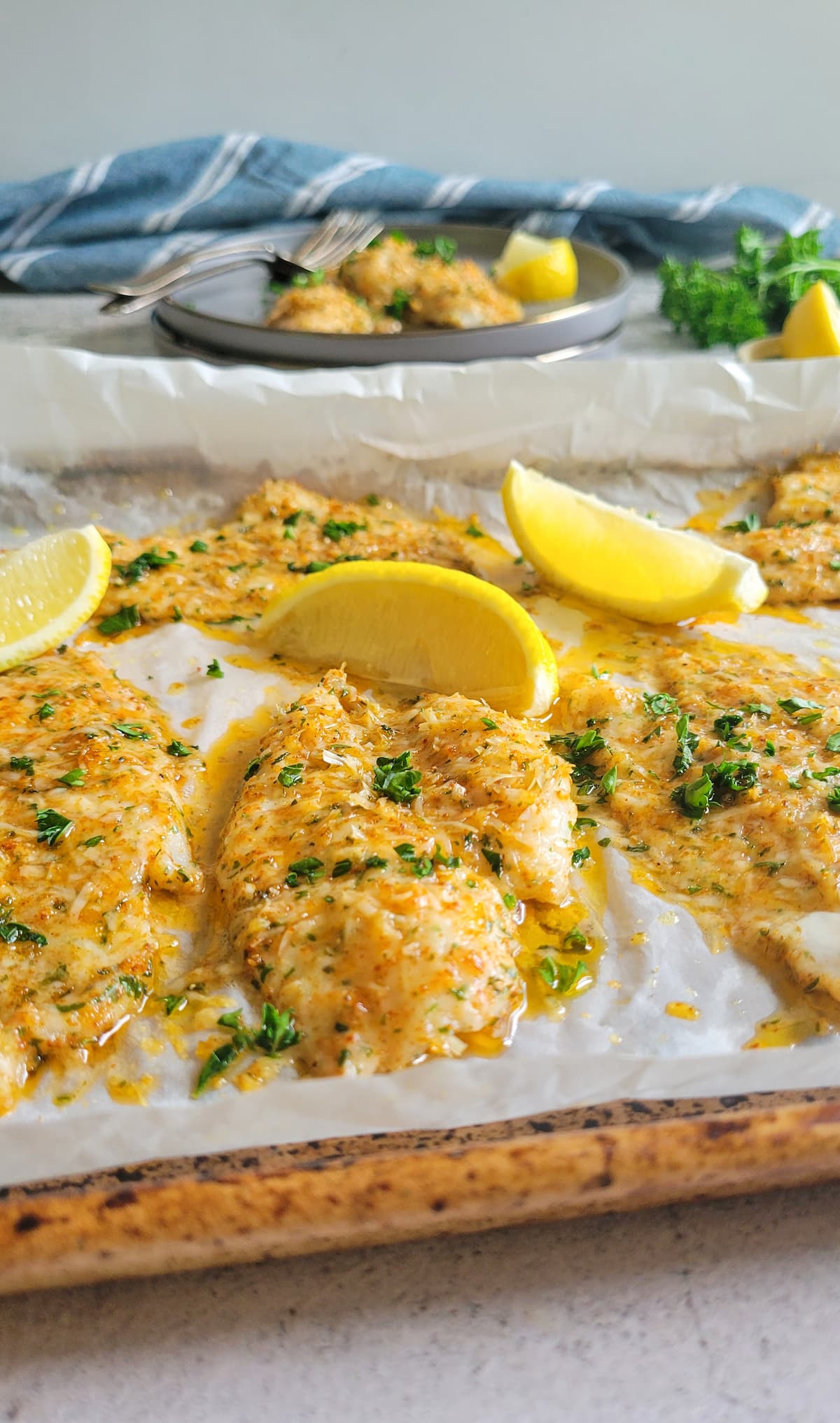 sheet pan of parmesan crusted fish garnished with fresh chopped parsley and lemon wedges, plate with some fish and lemon on it in the background