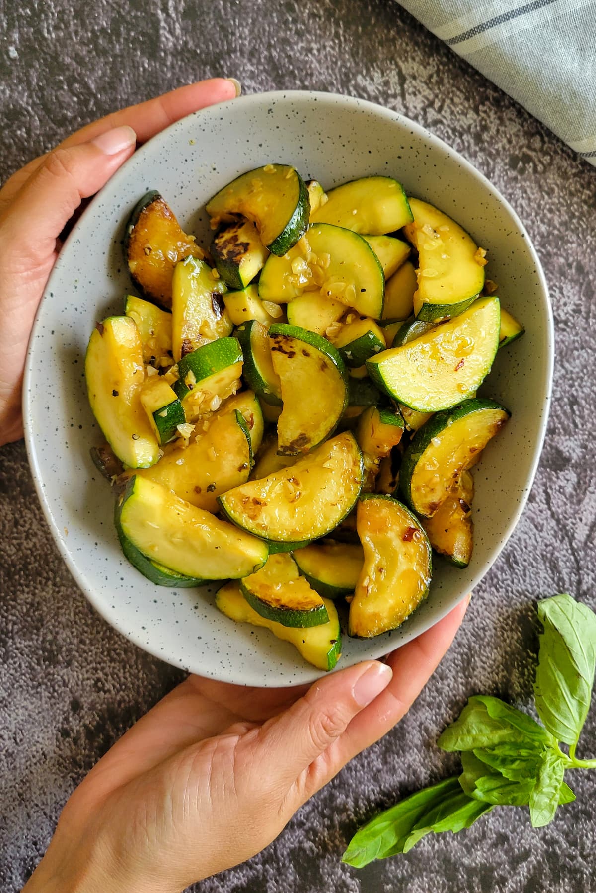 hands holding a bowl of sauteed zucchini with garlic, sprig of fresh basil on the side