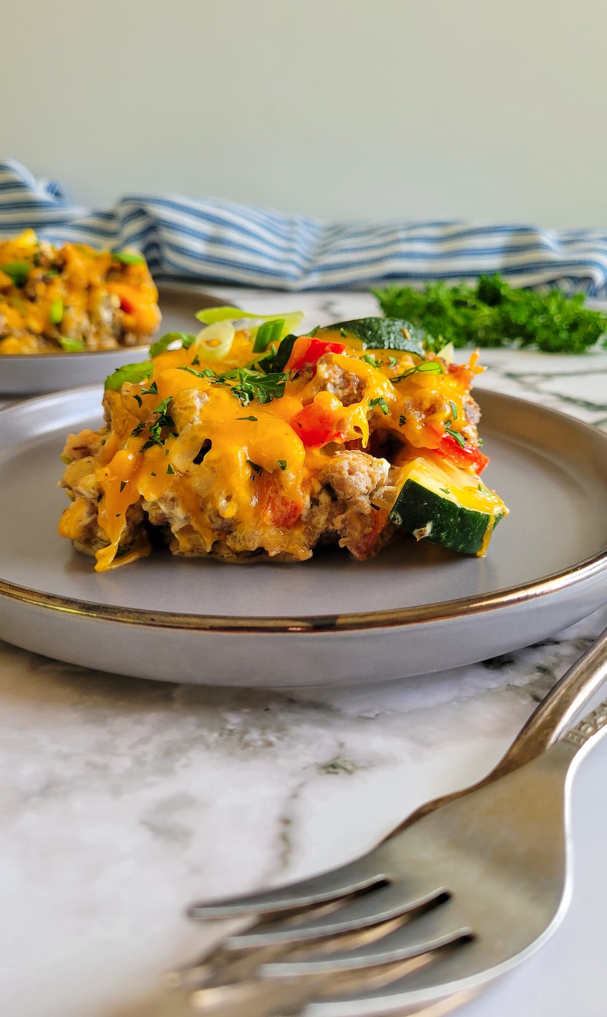 slice of sausage casserole with zucchini, melted cheddar and other veggies on a plate, another plate of it in the background