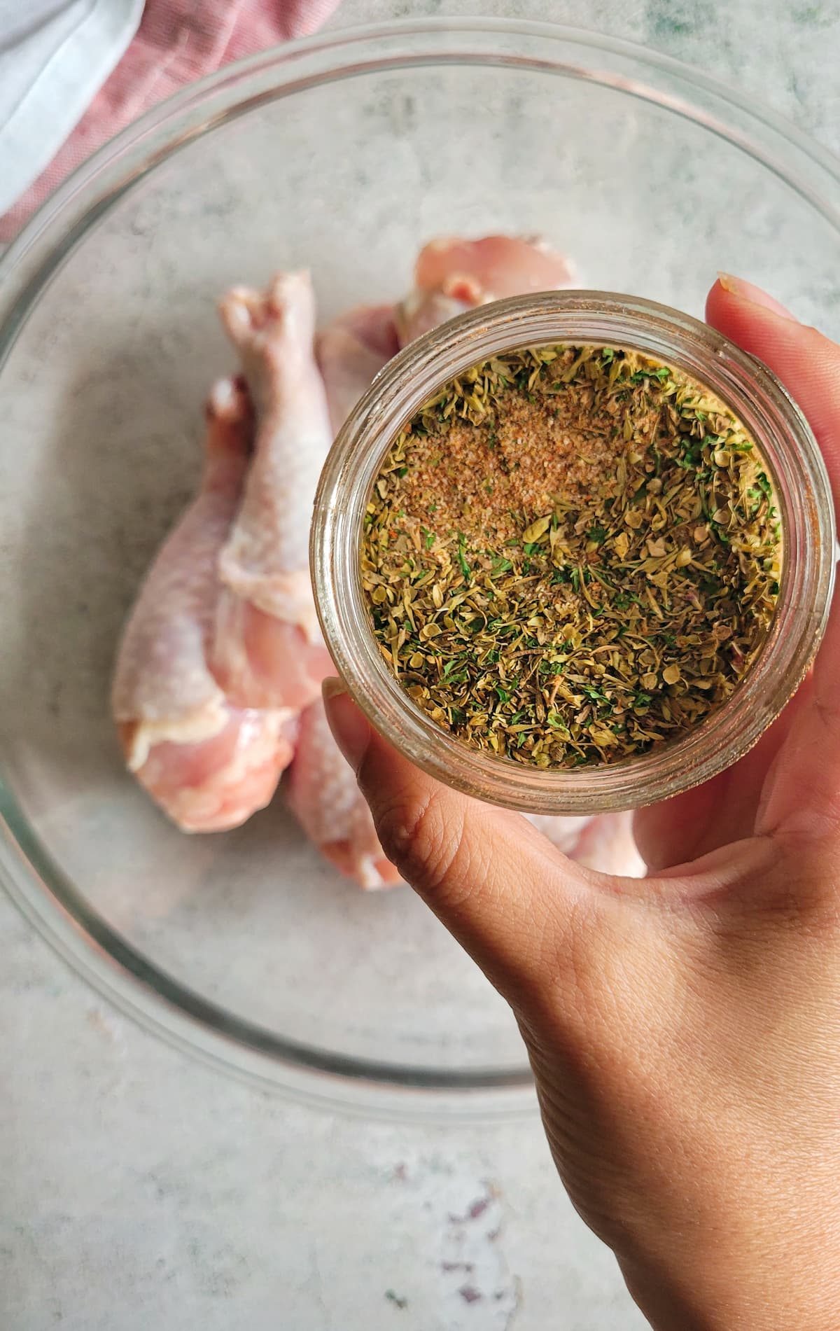 hand holding a jar of spices over a bowl of raw chicken drumsticks