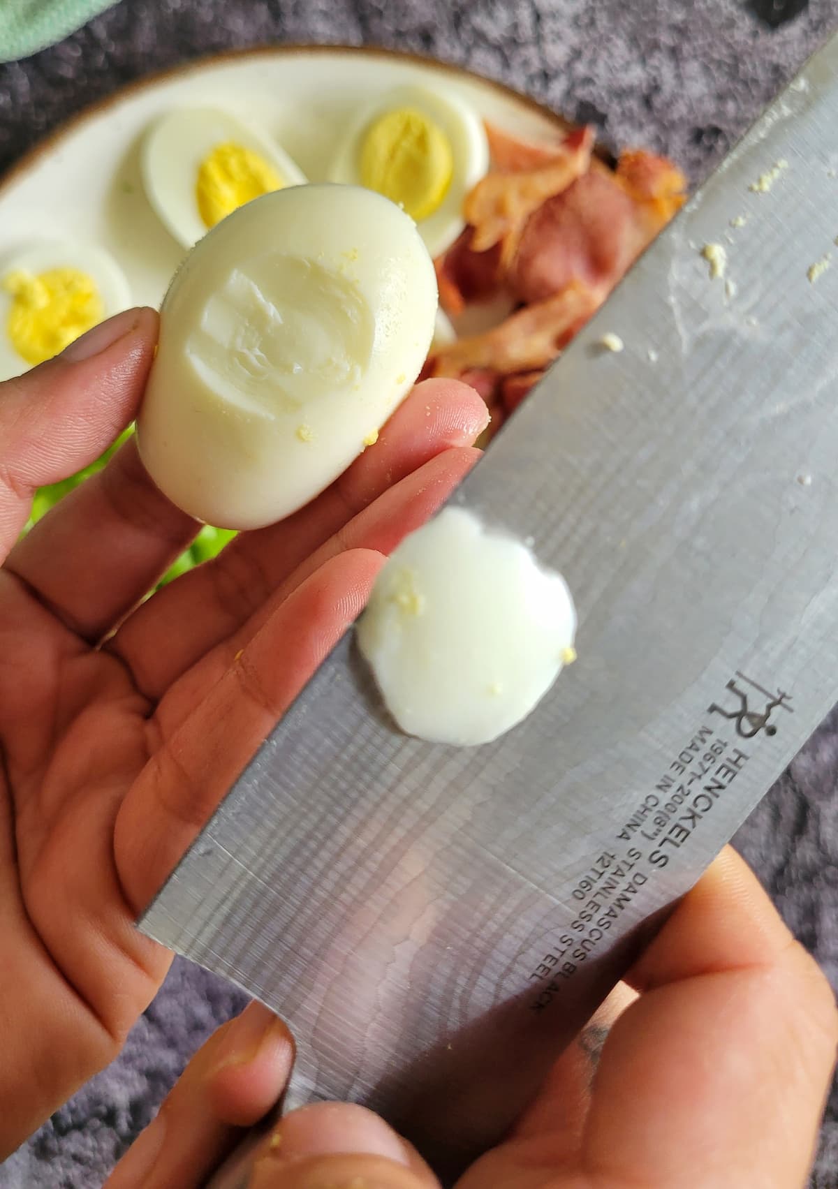 hand with a knife in one hand and a sliced hard boiled egg in the other, slice of the egg on the knife