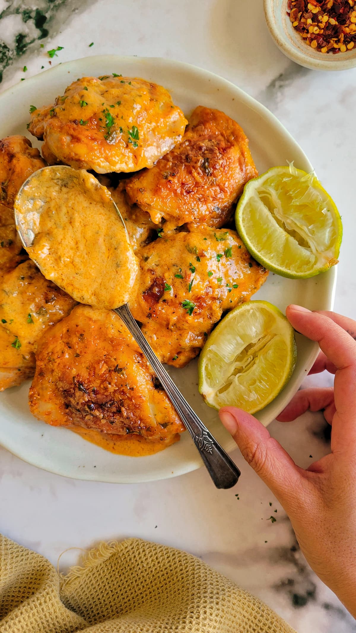 chicken thighs in orange sauce on a plate with a spoon and two limes, hand grabbing a lime