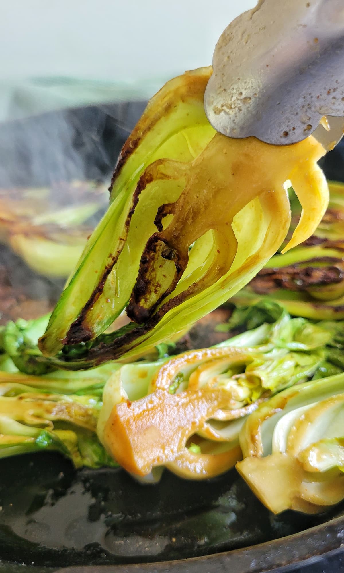 tongs lifting up a piece of caramelized bok choy from a cast iron skillet