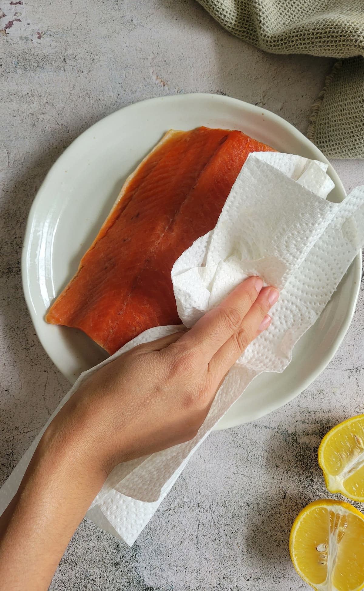 hand with a paper towel patting a piece of raw salmon dry, cut lemon in the background