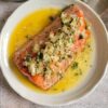 large piece of salmon on a plate in melted butter with diced cooked onions and chopped parsley