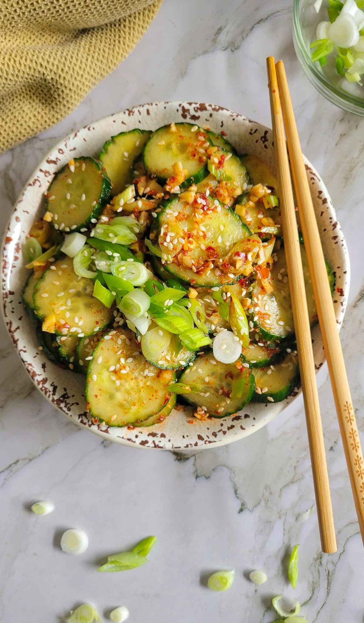 a bowl of cucumbers in a spicy sauce with sesame seeds and green onions, chopsticks on the bowl
