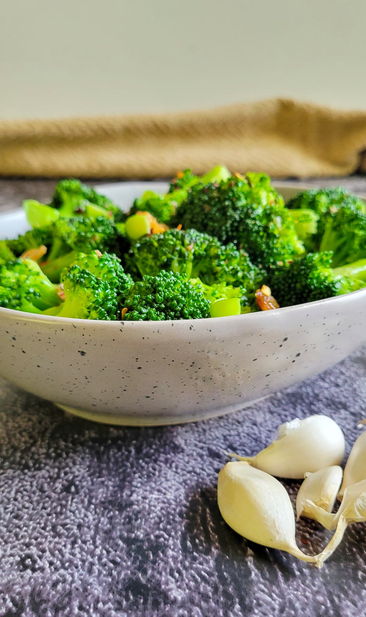 side view of a bowl of broccoli, unpeeled garlic cloves in front of the bowl