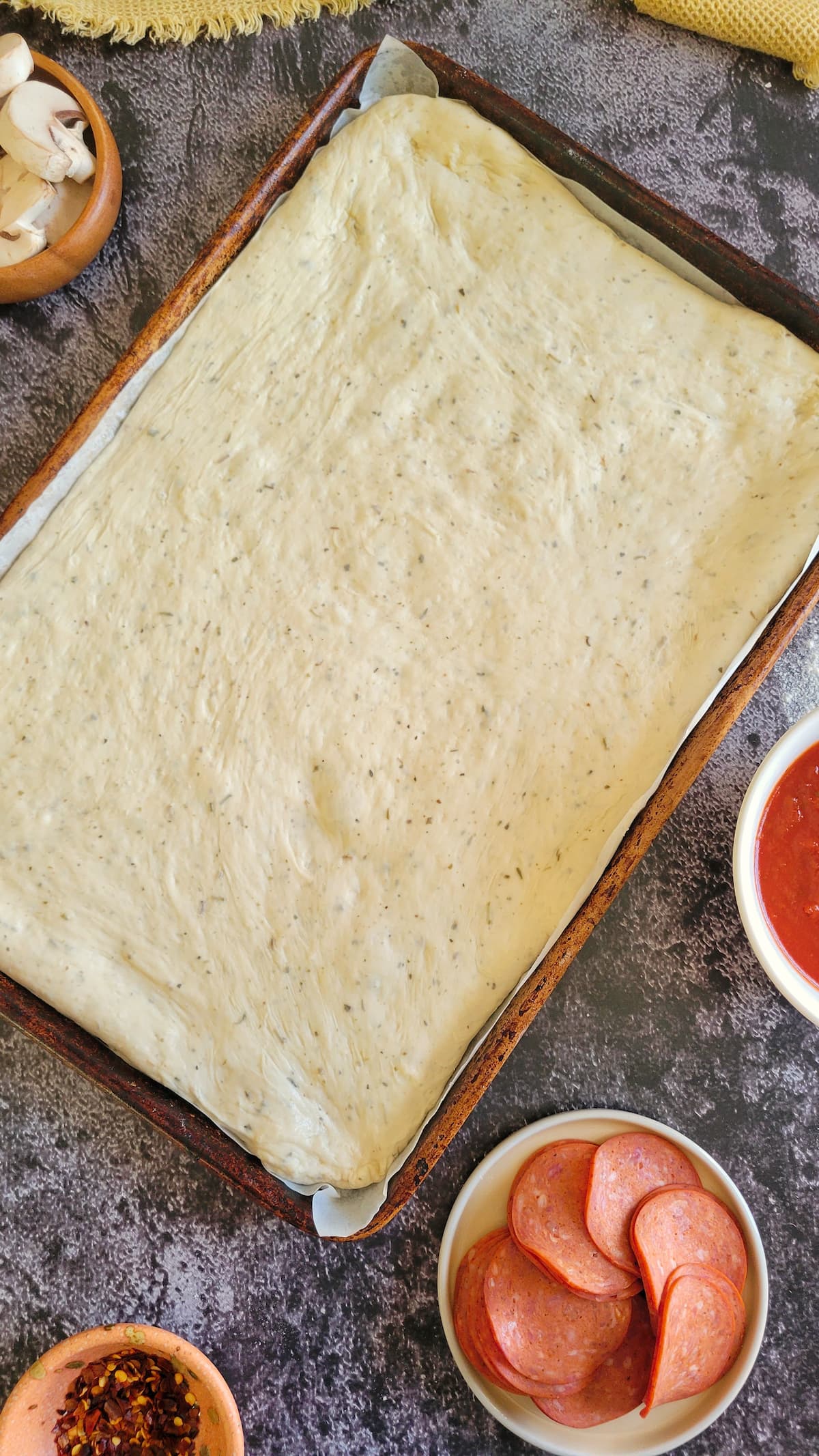 raw pizza dough on a parchment lined baking sheet surround by bowls of pizza toppings