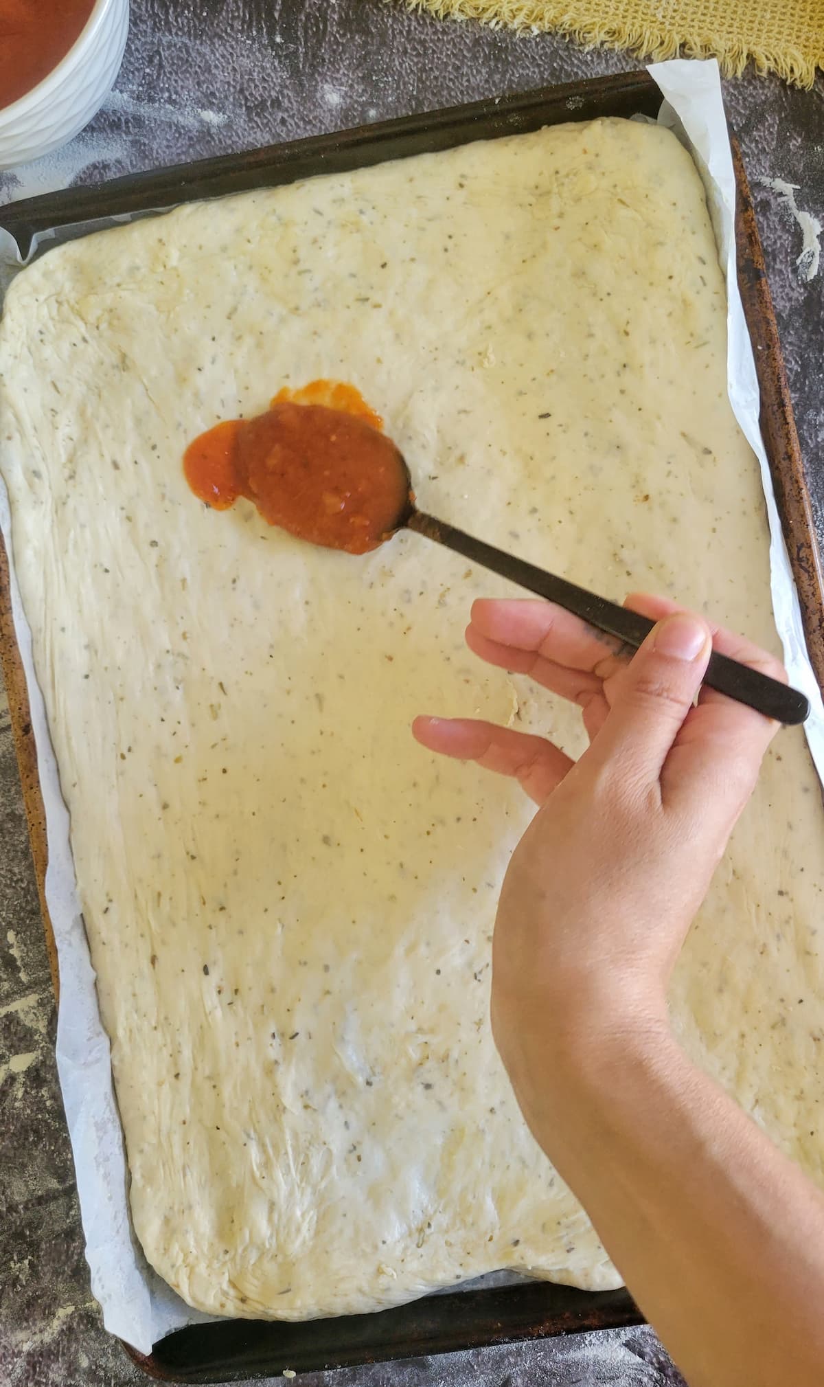 hand spooning some tomato sauce onto raw pizza crust in a parchment lined baking sheet
