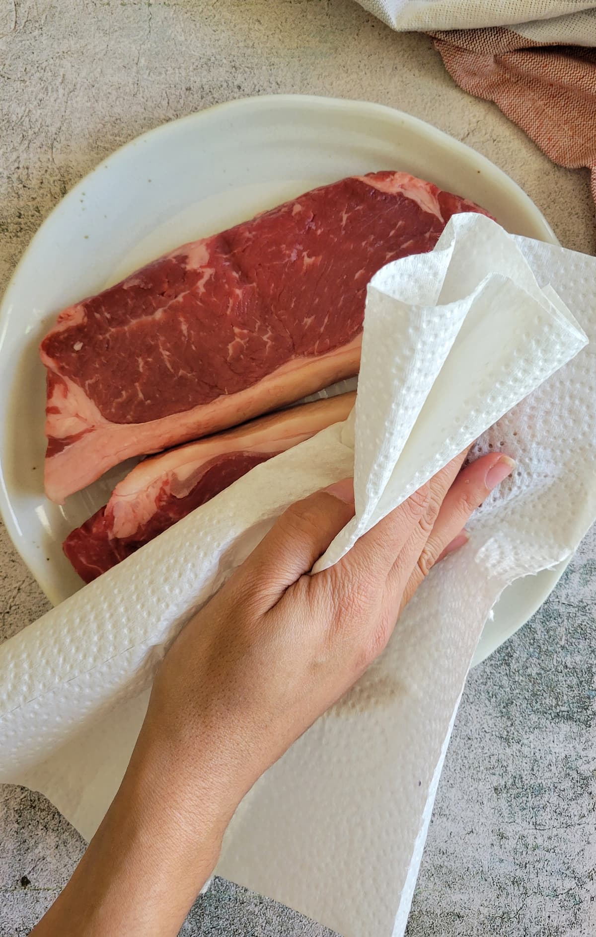 hand with a paper towel patting down two raw steaks on a plate