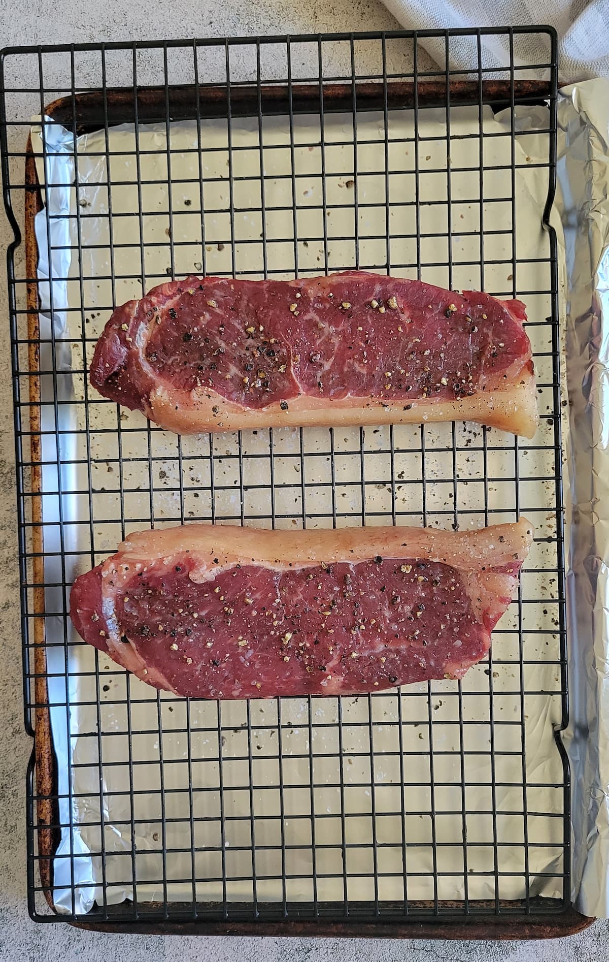 two salt and pepper seasoned steaks on a wire rack on an aluminum foil lined baking sheet