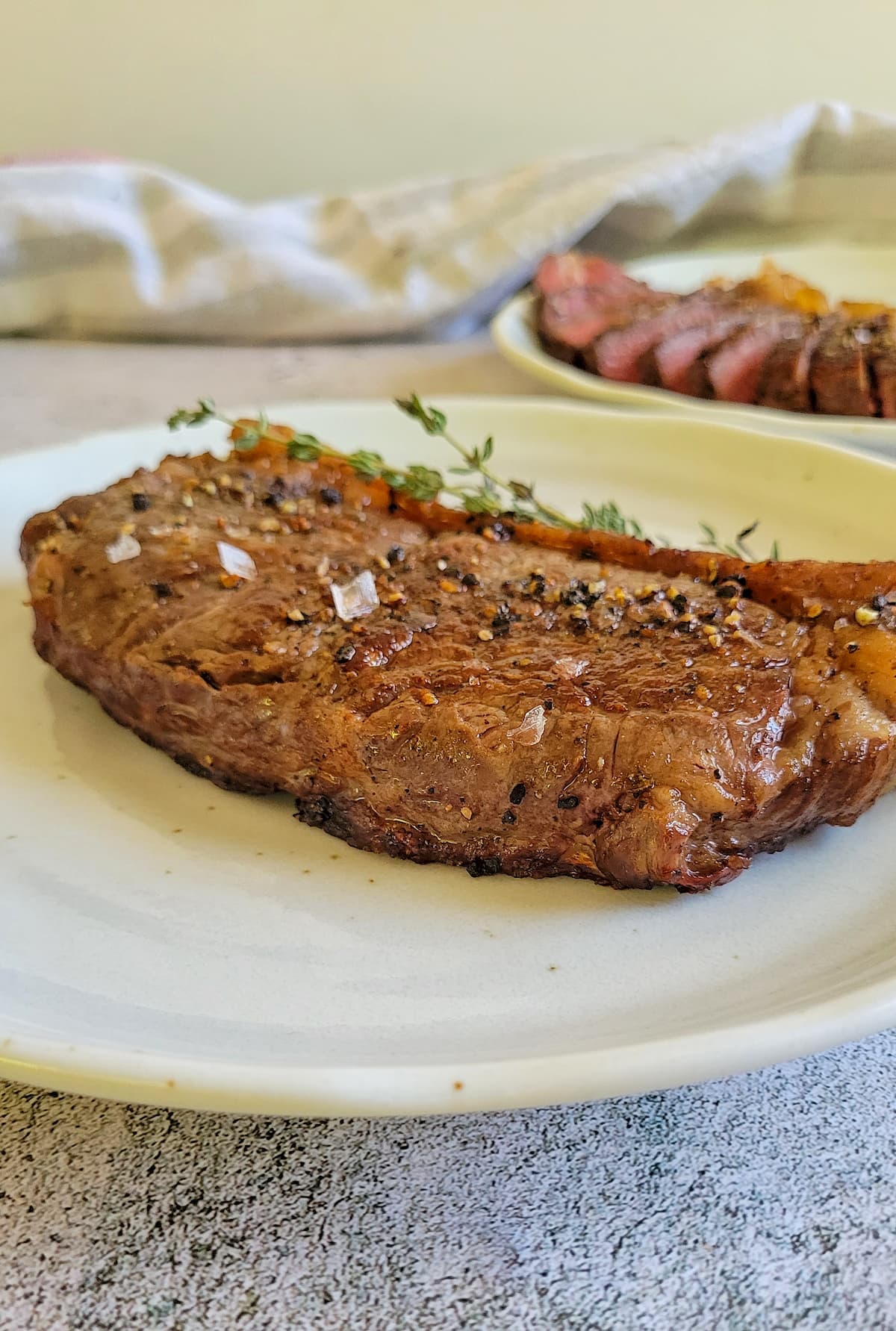 whole steak on a plate garnished with salt and pepper with a sliced on in the background