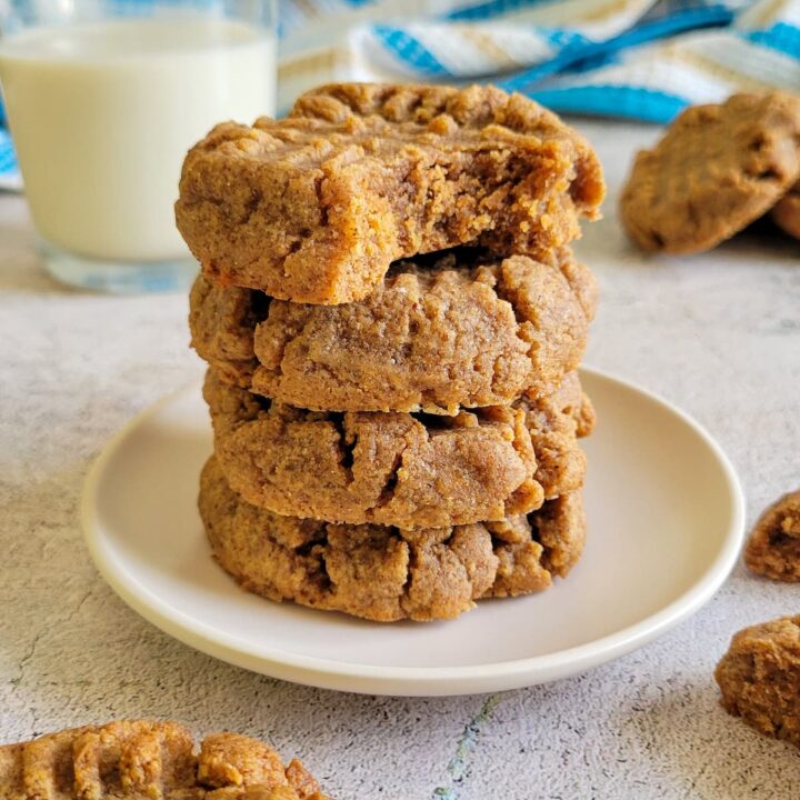 4 cookies stacked on a plate, top one has a bite taken out of it, more cookies and a glass of milk in the background