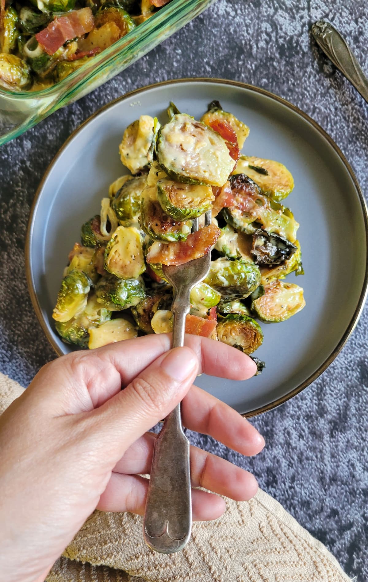 fork holding some brussels sprouts with bacon in a creamy sauce over a plate and dish with the rest of it