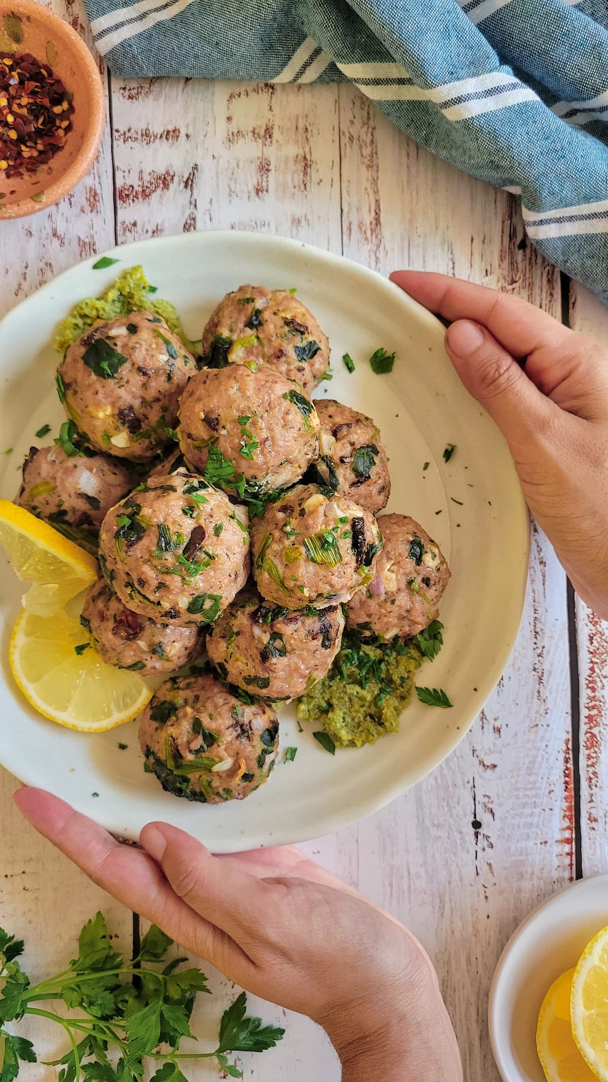 hands around a plate of meatballs on pesto, garnished with fresh chopped parsley and lemon