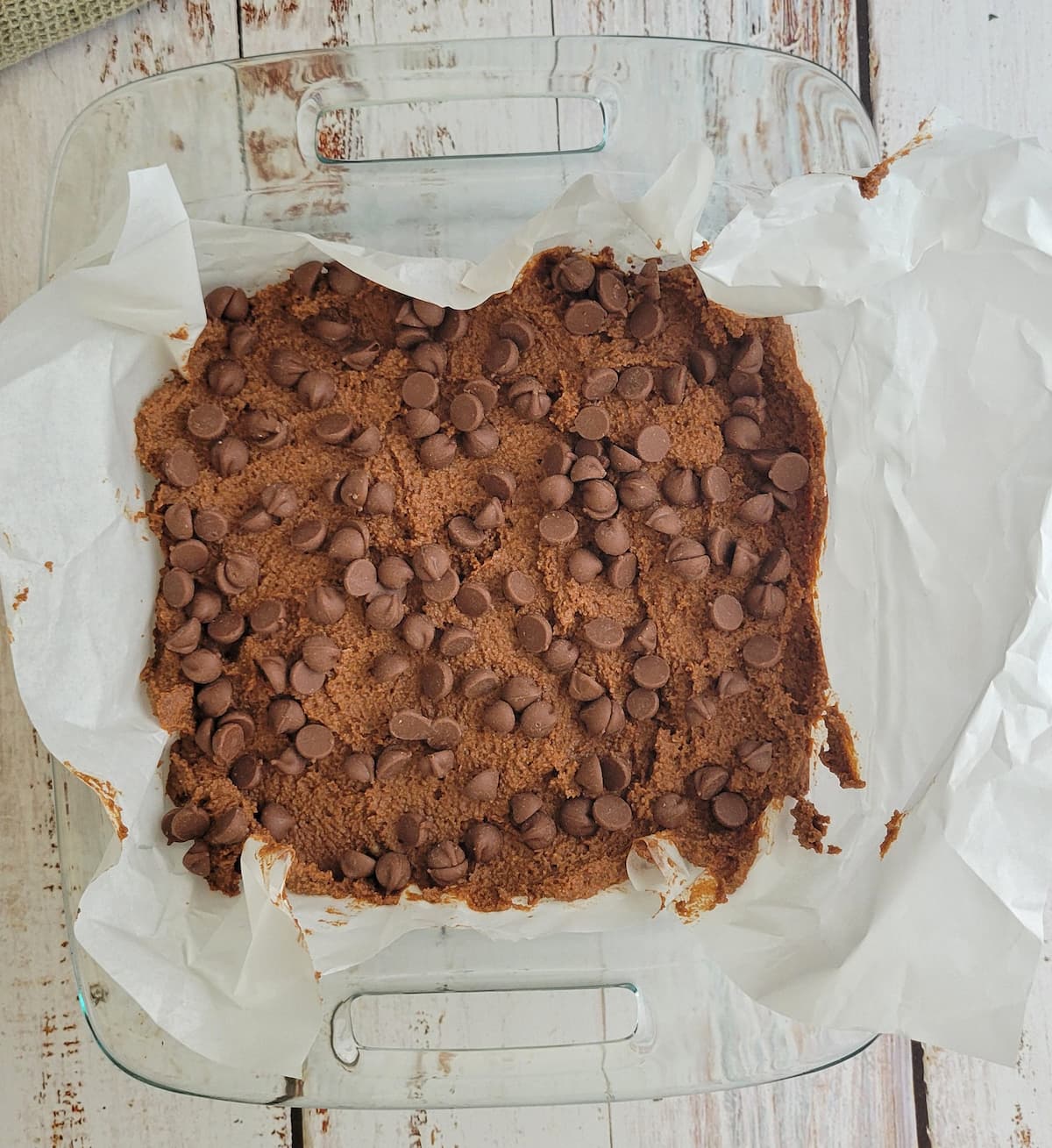 brownie batter topped with chocolate chips in a baking dish lined with parchment paper