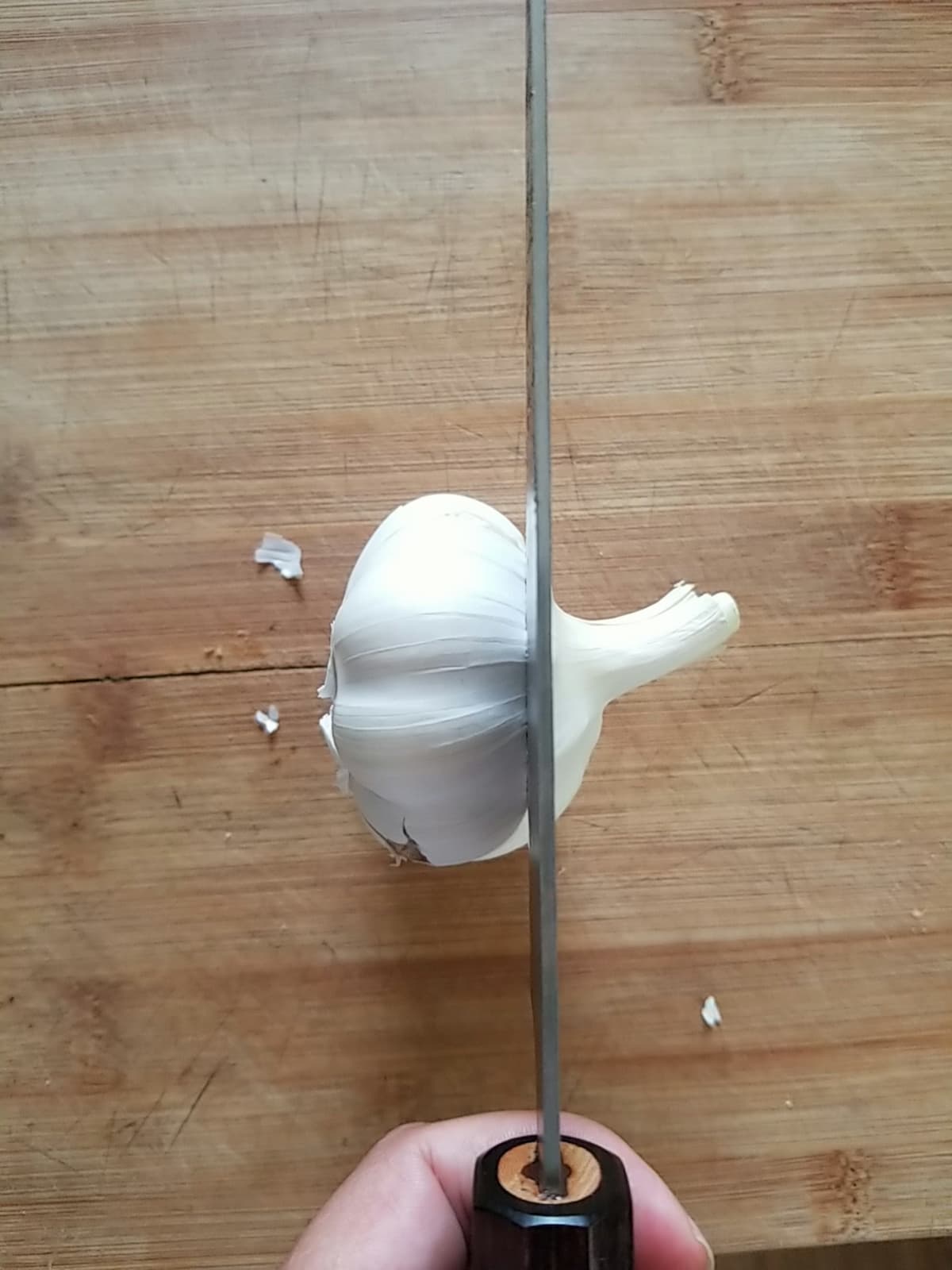 knife cutting off 1/4-inch off the top of a head of garlic on a cutting board