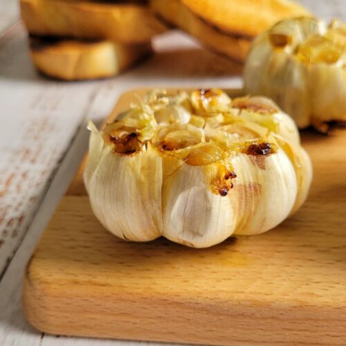 roasted garlic bulb on a cutting board, toasted bread and more roasted garlic in the background