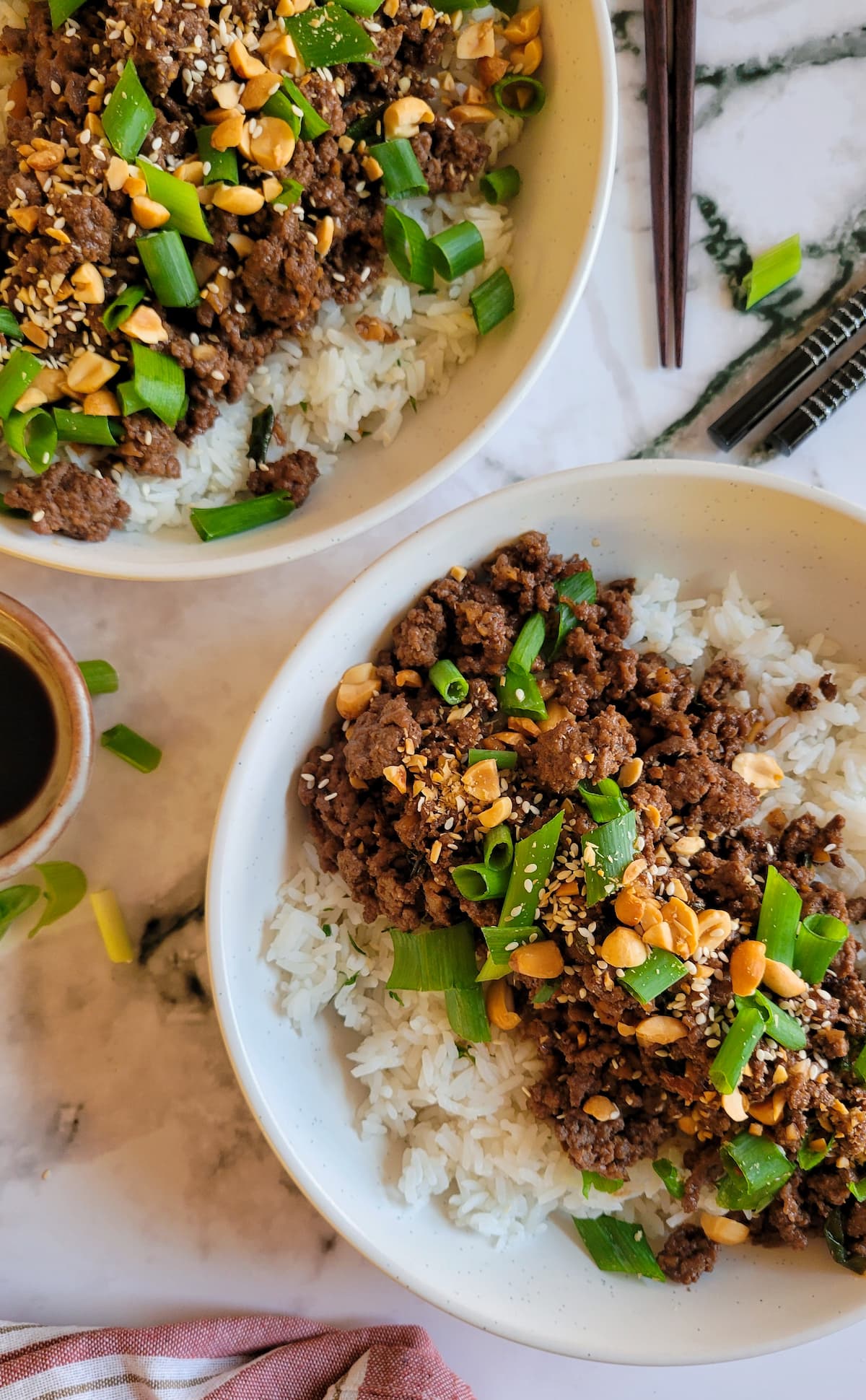 two ground beef and rice bowls with chopped peanuts, sesame seeds and green onions, chopsticks in the background