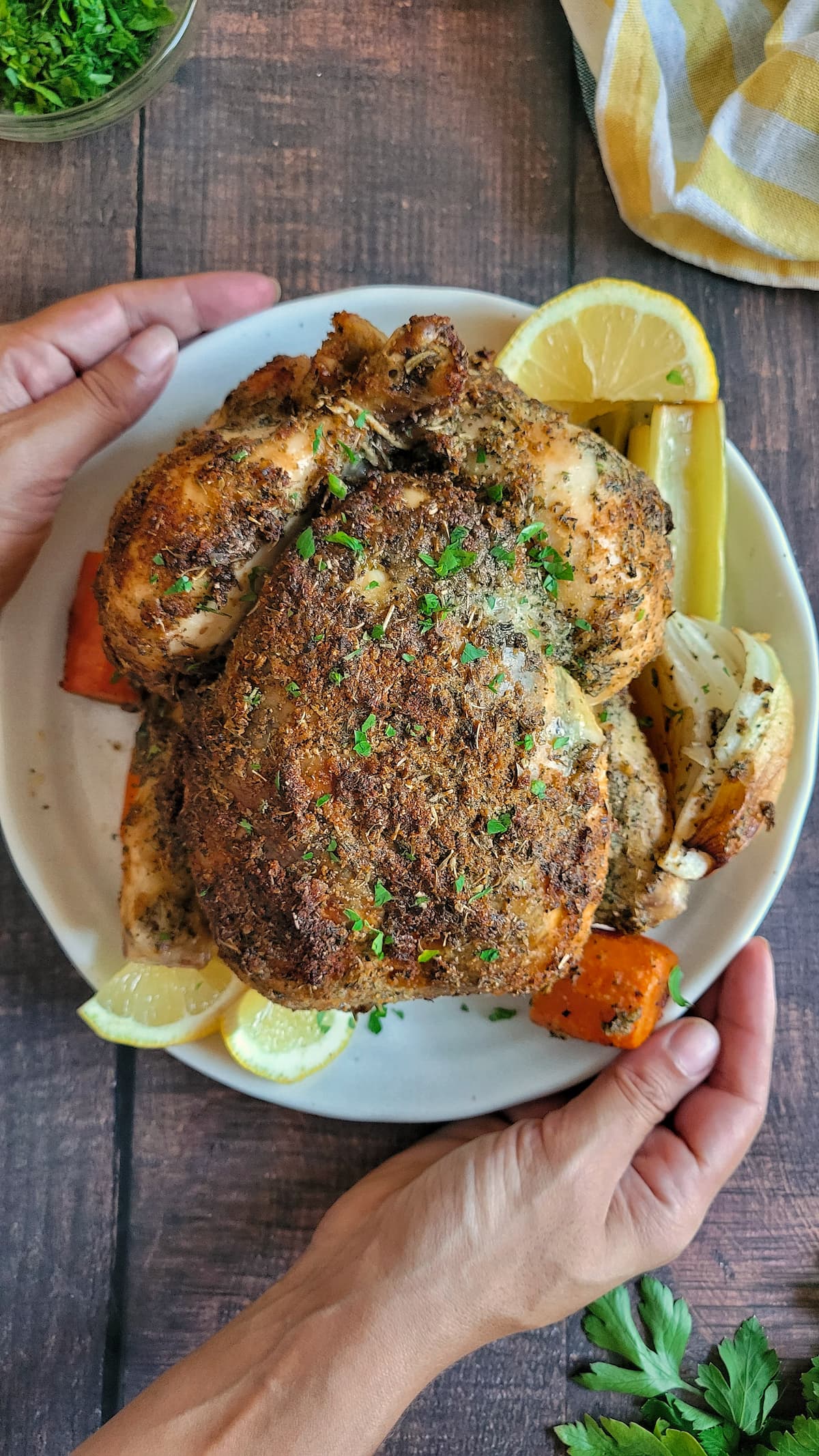 hands holding a plate with a whole roasted chicken with lemon, herbs and carrots