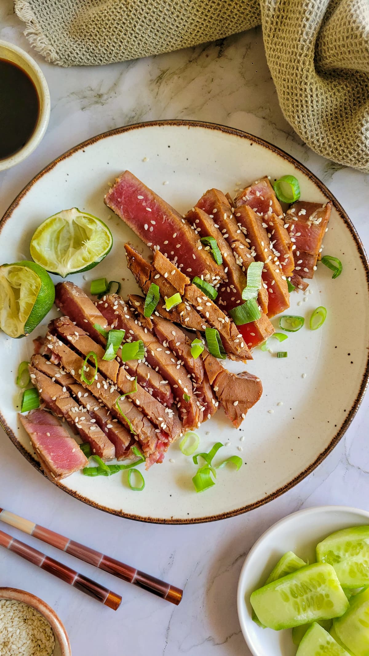 sliced rare tuna steak on a plate with green onions, sesame seeds, lime, cucumber, chopsticks, soy sauce and more sesame seeds in the background