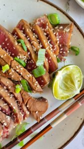 sliced rare tuna steak on a plate with green onions, sesame seeds, lime and chopsticks.