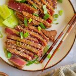 sliced rare tuna steak on a plate with green onions, sesame seeds, and lime with chopsticks, soy sauce and more sesame seeds in the background.