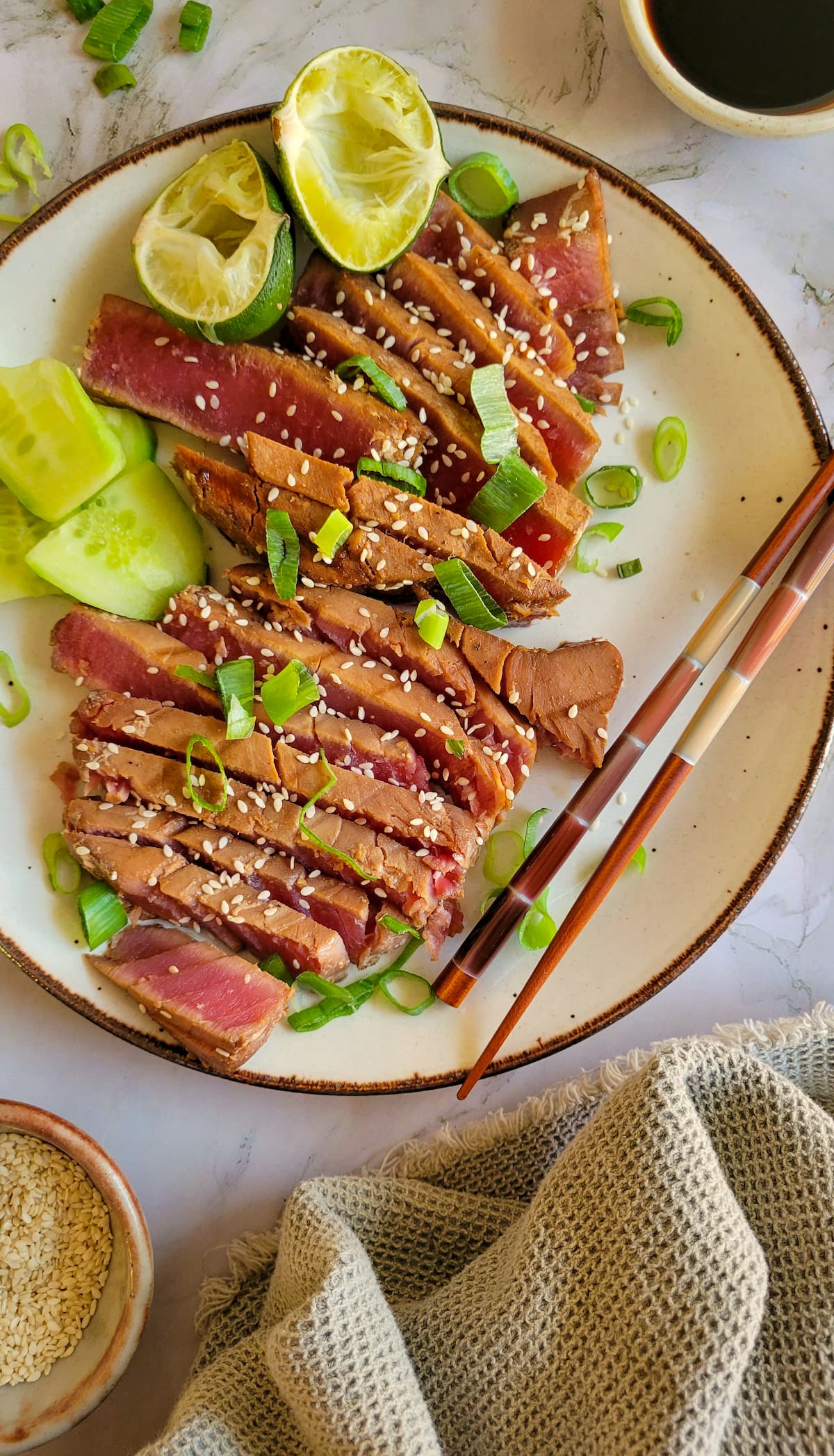 sliced rare tuna steak on a plate with green onions, sesame seeds, lime with chopsticks, soy sauce and more sesame seeds in the background