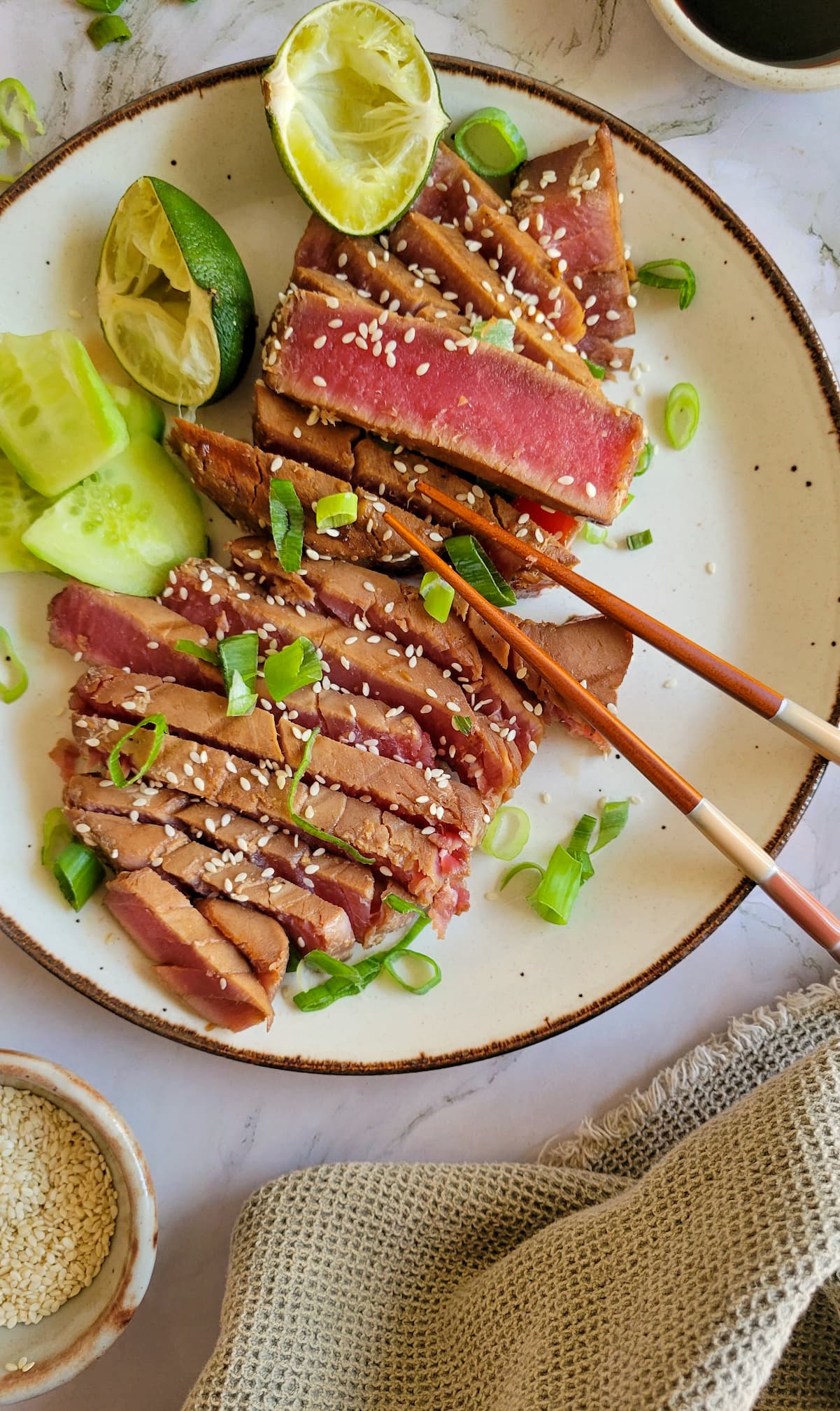 sliced rare tuna steak on a plate with green onions, sesame seeds, lime, cucumber and chopsticks