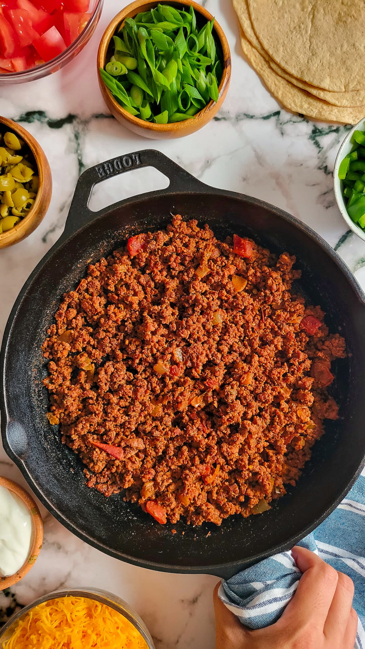 hand holding a skillet with taco seasoned ground beef, bowls of chopped veggies surrounding it