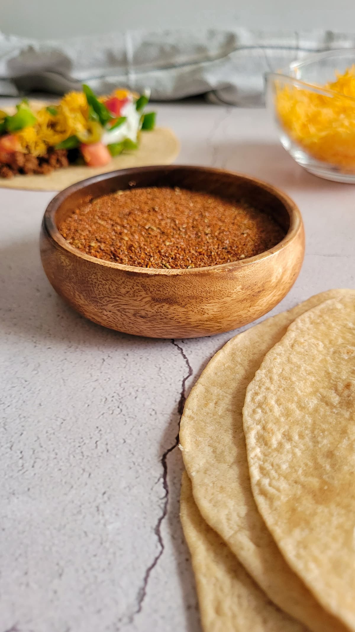 taco seasoning mix in a bowl, flour tortilla, bowl of cheese and a fully loaded taco in the background