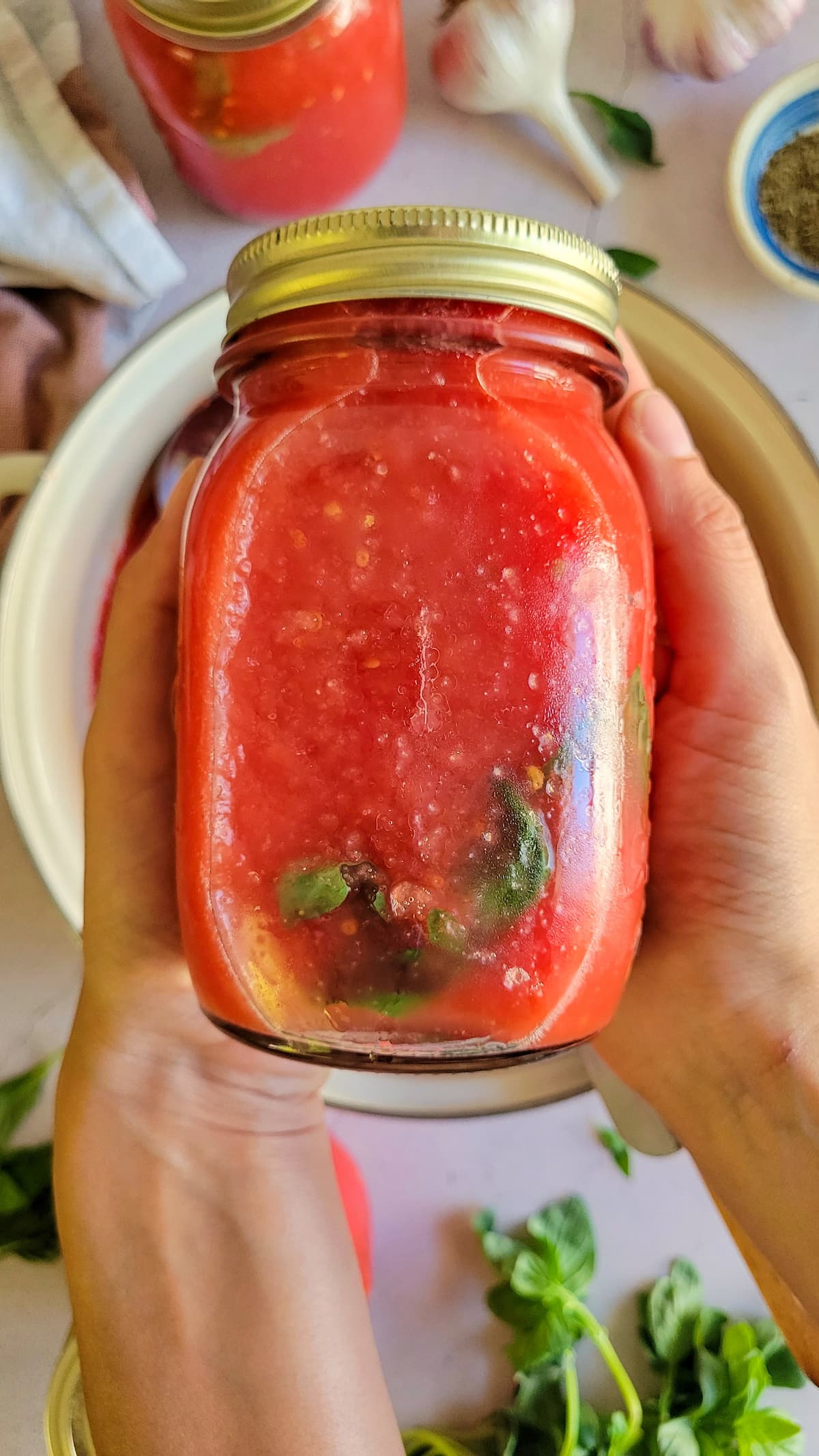 hands holding a jar of tomato sauce with basil, more herbs and jars in the background