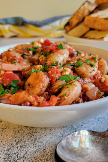 side view of a bowl of shrimp with tomato sauce, feta and fresh herbs, plate of toasted bread in the background
