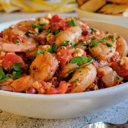 side view of a bowl of shrimp with tomato sauce, feta and fresh herbs, plate of toasted bread in the background
