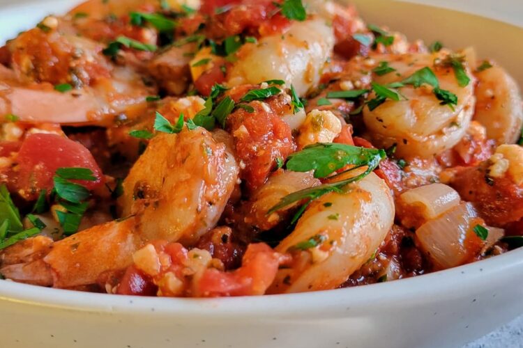 side view of a bowl of shrimp with tomato sauce, feta and fresh herbs, plate of toasted bread in the background