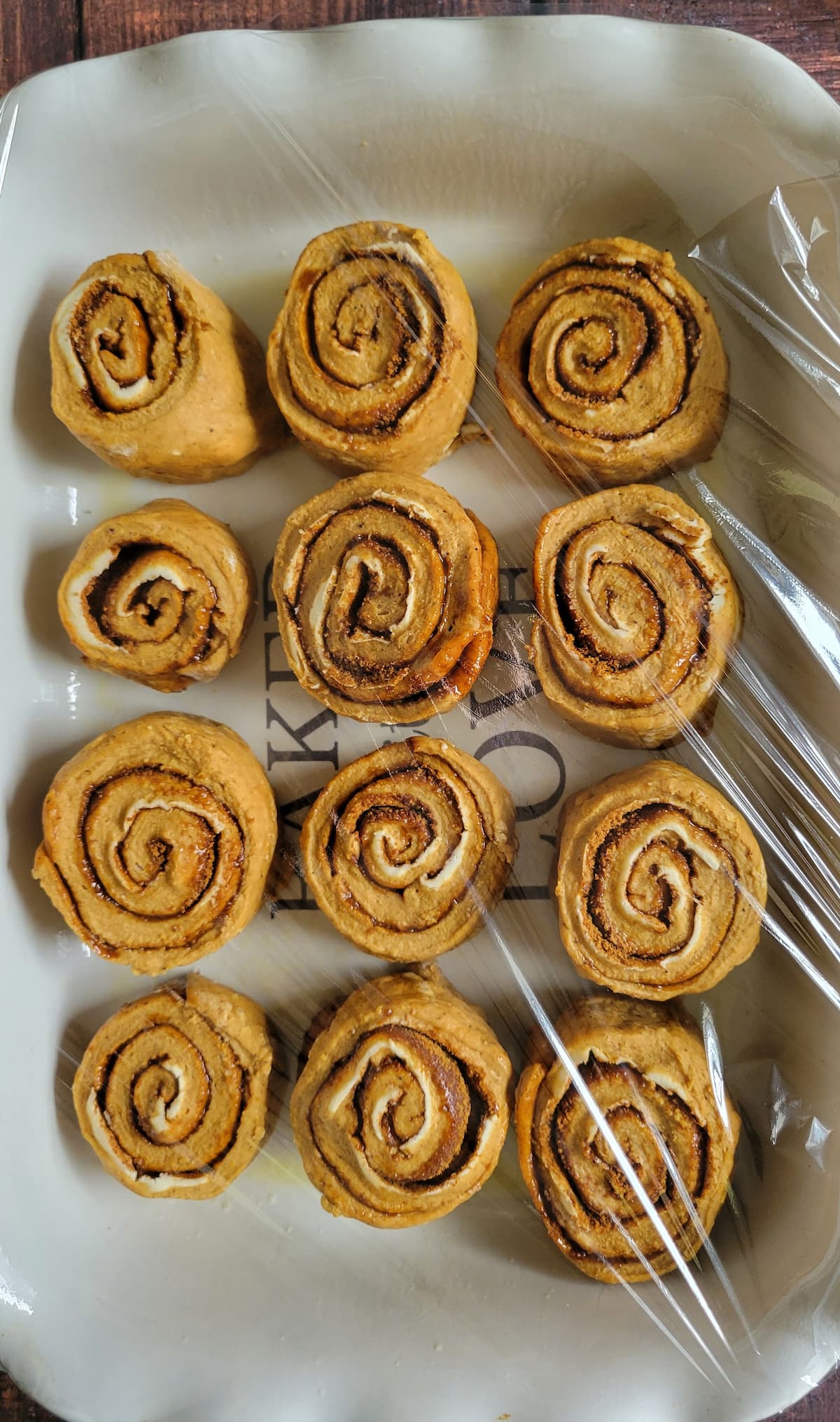 12 unbaked pumpkin cinnamon rolls in a tray with a piece of siran wrap over top