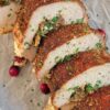 sliced stuffed pork tenderloin on a parchment lined baking sheet with fresh chopped parsley, goat cheese, fresh cranberries and caramelized onions
