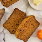 two slices of banana bread next to a whole half loaf and plate of butter