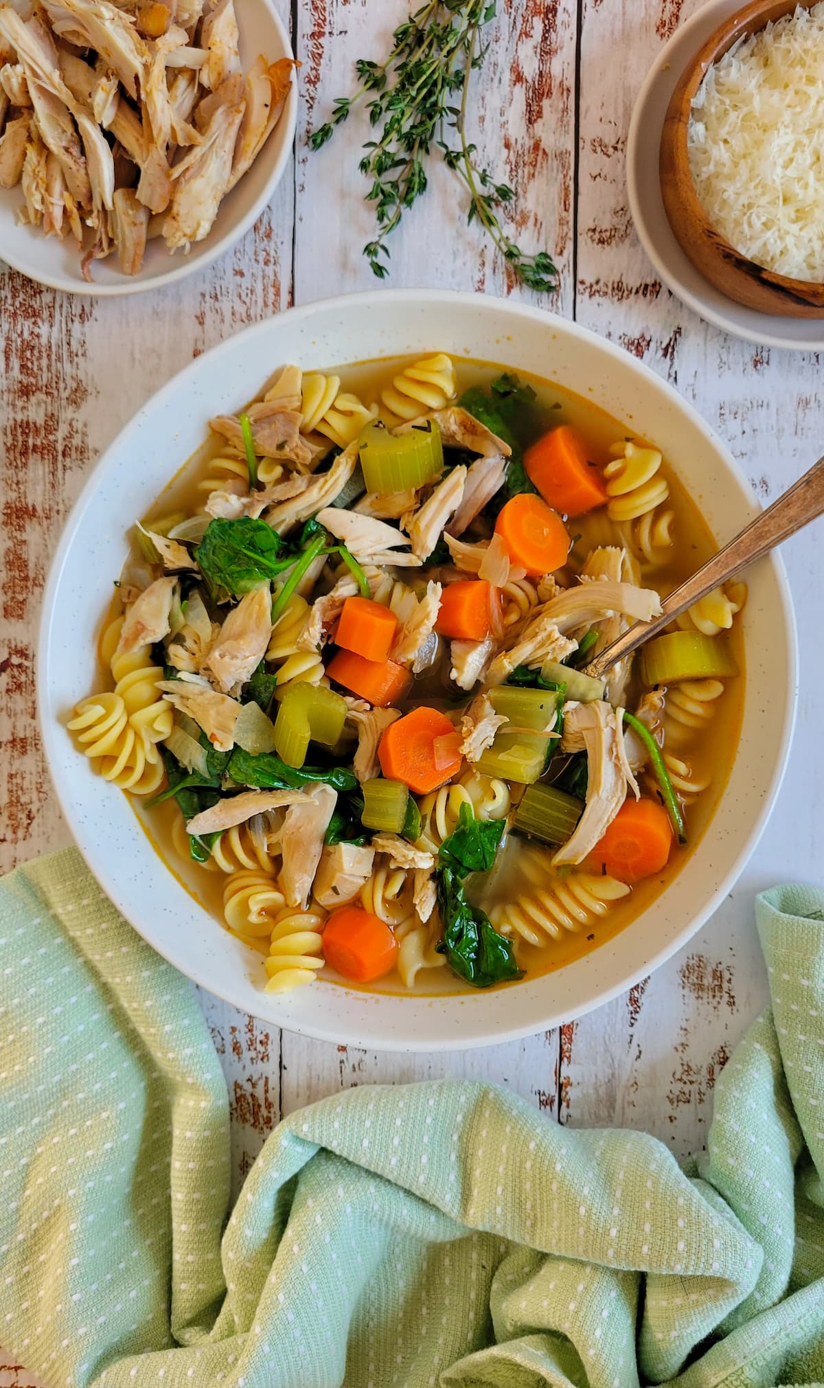 bowl of soup with turkey, vegetables and pasta, spoon in the bowl, plate of shredded turkey and parmesan in the background