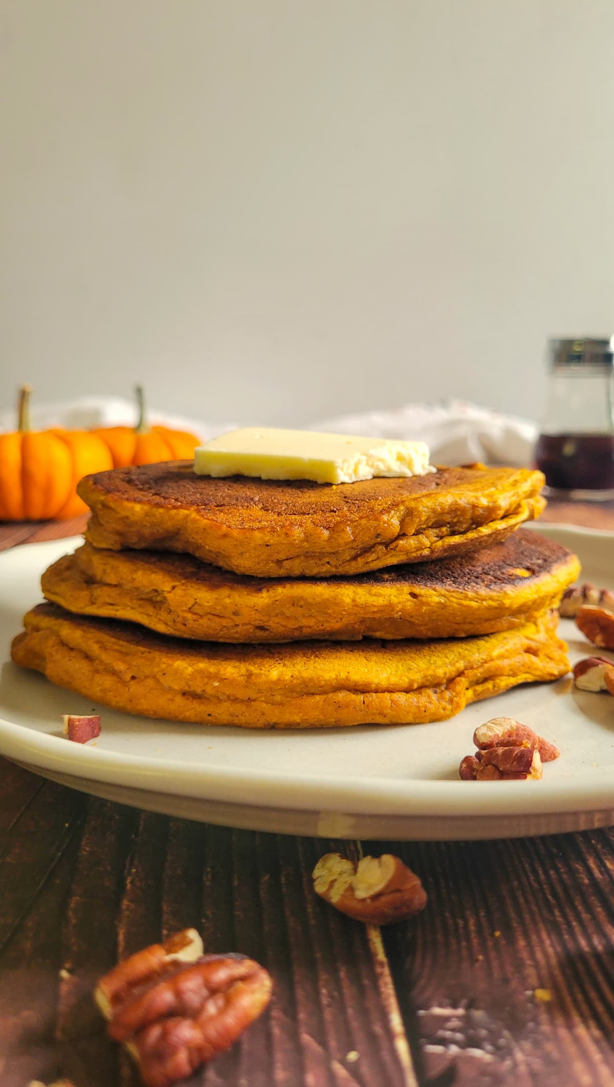 3 pancakes stacked on a plate with pecans and a pat of butter, pumpkins and syrup in the background